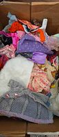 Open box showing large pile of varied Barbie garments including faux fur coat, knit jacket, sequined purple top and multicolored fabrics