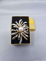 Front view of gold tone floral brooch showing rhinestones and central faux pearl in presentation box