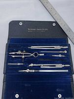 Open case displaying all eight metal drafting instruments against blue velvet lining