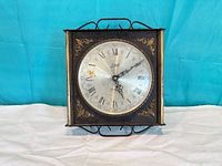 Front view of square metal clock showing dial, frame, hands and decorative accents