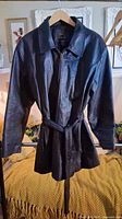 Full view of black leather trench coat on hanger, belt tied at waist