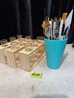 Cup containing assorted artist brushes and palette knives alongside wooden rack with glass vials
