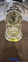 Front view of gold-tone anniversary clock under glass dome showing dial and pendulum