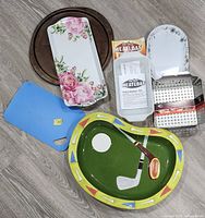 All items spread on floor showing full lot: golf serving bowl, floral tray, meatloaf pan with insert, grill pan, cutting board, nested pizza pans