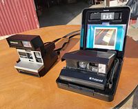 Both Polaroid cameras on table: Spectra System SE in case and Sun 600 LMS