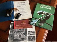 All Jaguar memorabilia items arranged: service manual binder, Great Marques book, key with fob, aviator sunglasses, maintenance vouchers booklet, promotional brochure