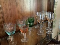 Group of twelve vintage aperitif glasses displayed on glass shelf: six clear stemmed glasses to the right, six colored spiral-stem glasses to the left