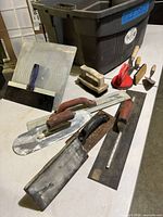 Tile tools laid out: trowels, floats, suction lifter, wire brush in front of tote