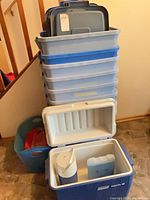 Stack of 8 blue and clear plastic bins with lids, cooler open showing contents, misc bin visible
