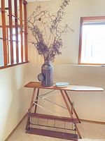 Overall view of ironing board with attached roll holder, metal canister holding dried flowers, and painted metal dish