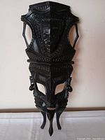 Front view of dark carved wooden tribal mask showing central face, layered figures, geometric patterns, elongated form