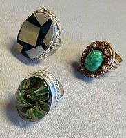 Three rings grouped: Avon poison ring (geometric inlay), gold-tone faux jade cluster ring, silver-tone green art glass ring