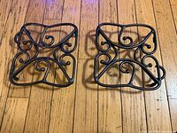 Two square wrought iron trivets with scrollwork on hardwood floor