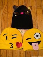 Three girls knit beanie hats laid out on hardwood floor: black cat-ear design, two yellow emoji designs