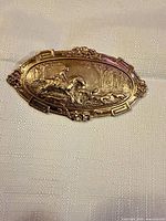 Front view of gold-tone repousse oval brooch with hunting scene relief