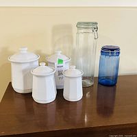 Four white ceramic canisters, clear glass clamp-lid jar, blue plastic jar on tabletop
