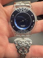 Front view of Swatch watch showing blue dial, crystal markers, scroll design, and ornate bracelet