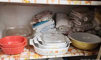 Shelf showing yellow Pyrex bowl, CorningWare casseroles, red stoneware bakers, glass lids and stacks of towels