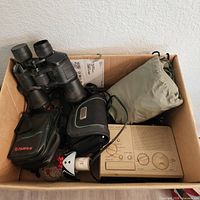 Overview of lot contents in box: binoculars, camera case, GPS case, radio, drawstring bag, ornament, adapter