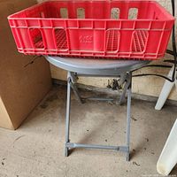 Round grey metal patio table with folding legs, red Coca-Cola plastic crate resting on top