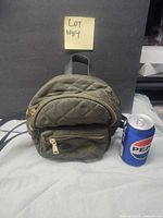 Front view of olive quilted mini backpack beside Pepsi can for scale