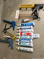 Top-down view showing five caulking guns and ten tubes of caulk laid out on concrete floor