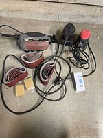 Two sanders with cords, sanding belts and pads on concrete floor