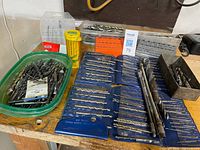 Wide view of assorted drill bits in organizer wallet, tray, tub and gauges on workbench