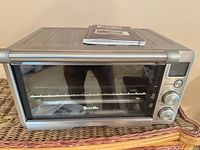 Front view of Breville compact toaster oven on wicker surface with wire rack inside and manual on top