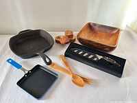 All items arranged: cast iron skillet, nonstick pan, wooden bowl, wooden spoon and fork, wooden triple dish, boxed chef’s knife