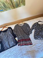 Three garments: black top with sheer panel, patterned dress with red hem, black floral blouse