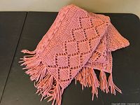 Folded mauve pink hand-knit afghan showing diamond and cable stitching with fringe ends