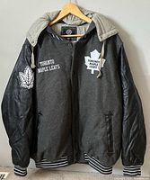 Front view of jacket on hanger showing wool body, leather sleeves, hood, embroidered logos, striped cuffs/hem