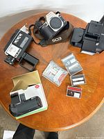 All items arranged on tabletop: Canon camera body, Minolta Super 8 camera, battery grip in box, flash with case, multiple cassette tapes