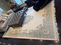 Full view of rug under furniture showing cream field, blue-green floral border, fringed edge