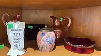 Shelf view showing two cranberry glass pitchers, hand-painted jar, compote and additional bowl