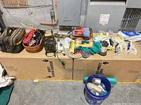 Wide view of all items on two cardboard boxes including tool bag, wicker basket, paint supplies, lanterns, bucket