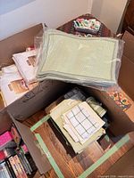 Boxes containing packaged white tablecloths, stack of green placemats in plastic, folded tea towels and other linens
