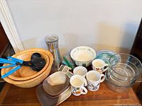 Group view of entire lot showing wooden bowls, glass bowls, mugs, utensils, carafe, plates, cutting board, timer
