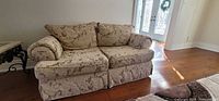 Front view of beige floral two-seat sofa showing cushions, rolled arms and skirted base