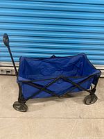 Side view of blue folding wagon showing fabric body, frame, wheels, and pull handle