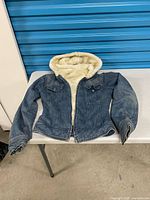 Front view of blue denim jacket with sherpa lining and hood displayed on table