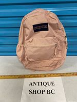 Front view of beige Jansport backpack showing logo patch, front pocket, mesh side pocket and overall wear