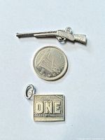 Rifle charm, coin for scale, and square "ONE" charm front view