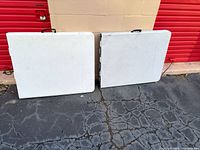 Two white folding tables folded in half, carry handles visible, standing upright on ground
