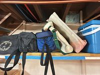 Shelf view showing black St. Luke’s insulated lunch bag, blue insulated lunch cooler, natural canvas tote, and blue hard plastic cooler