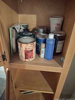 Overview of cabinet shelf with assorted paint, stain cans, grout tub, bottle and tools