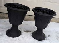 both black plastic pedestal urn planters side by side on snow
