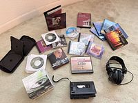 Assorted CDs, blank CD-R spindle, cassette player, headphones and storage cases spread on carpet