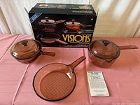 Boxed Visions cookware set displayed with two lidded saucepans, one skillet, booklet